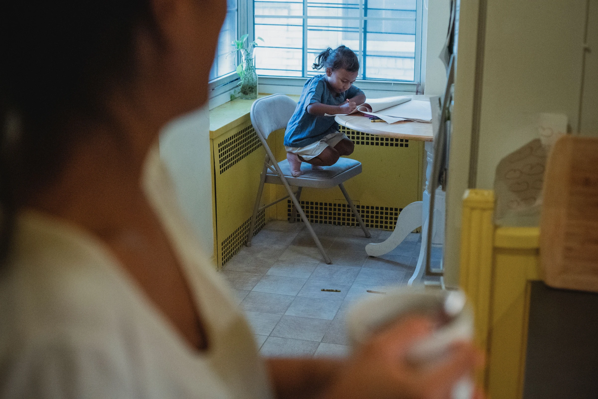 A child draws at a kitchen table while an adult watches from nearby.